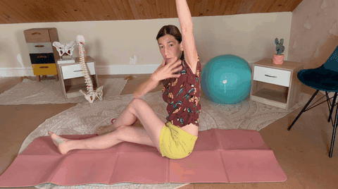 Une praticienne assise sur un tapis lève un bras au-dessus de la tête pendant un exercice de mobilité.